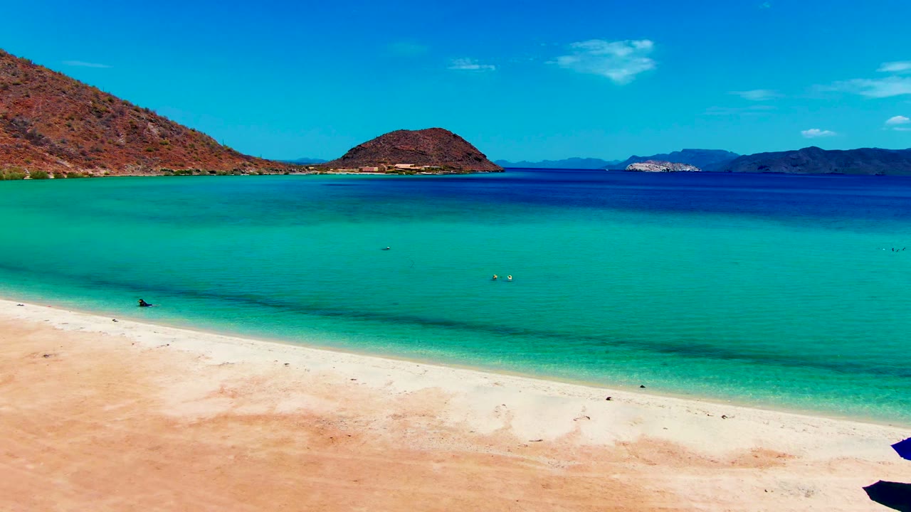 At Playa Santispac, the beach occupies the lower frame, clear green-blue shallows stretch outward, small swimmers sit near center right, and dry hills line the horizon.