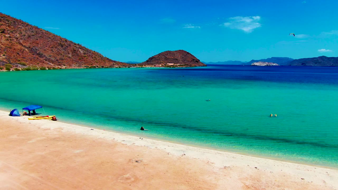 At 0:02, the Playa Santispac beach view shows the umbrella and gear farther inside the left foreground, swimmers as small figures in the water, and a broad turquoise-to-blue water gradient leading to hills and islands.