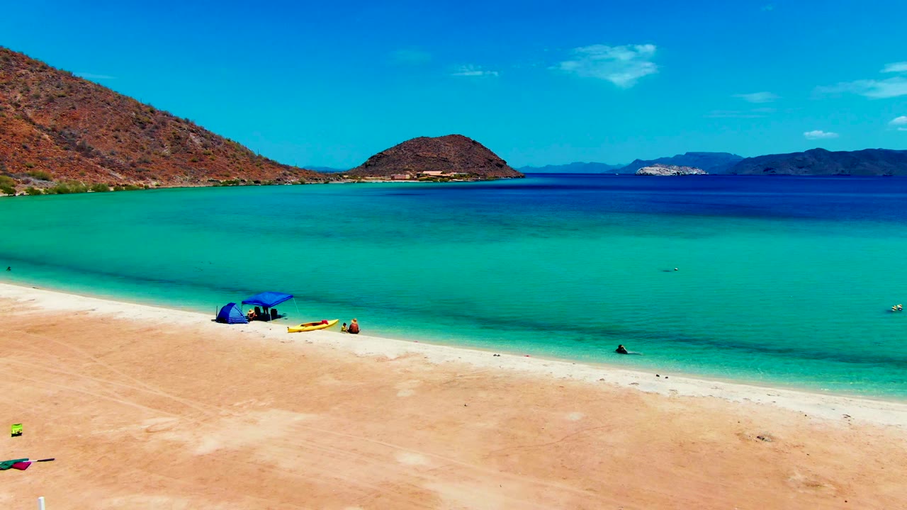 Opening view of Playa Santispac with pale sand, clear turquoise water, a blue umbrella near the shoreline, small people visible, and brown hills across the bay.