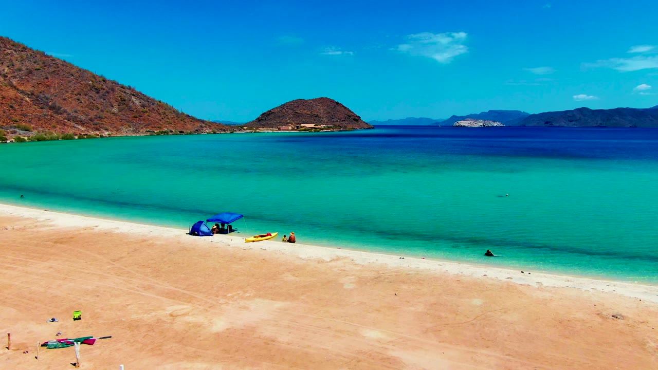 Sandy shoreline with turquoise shallows, beach umbrella, people, and hills.