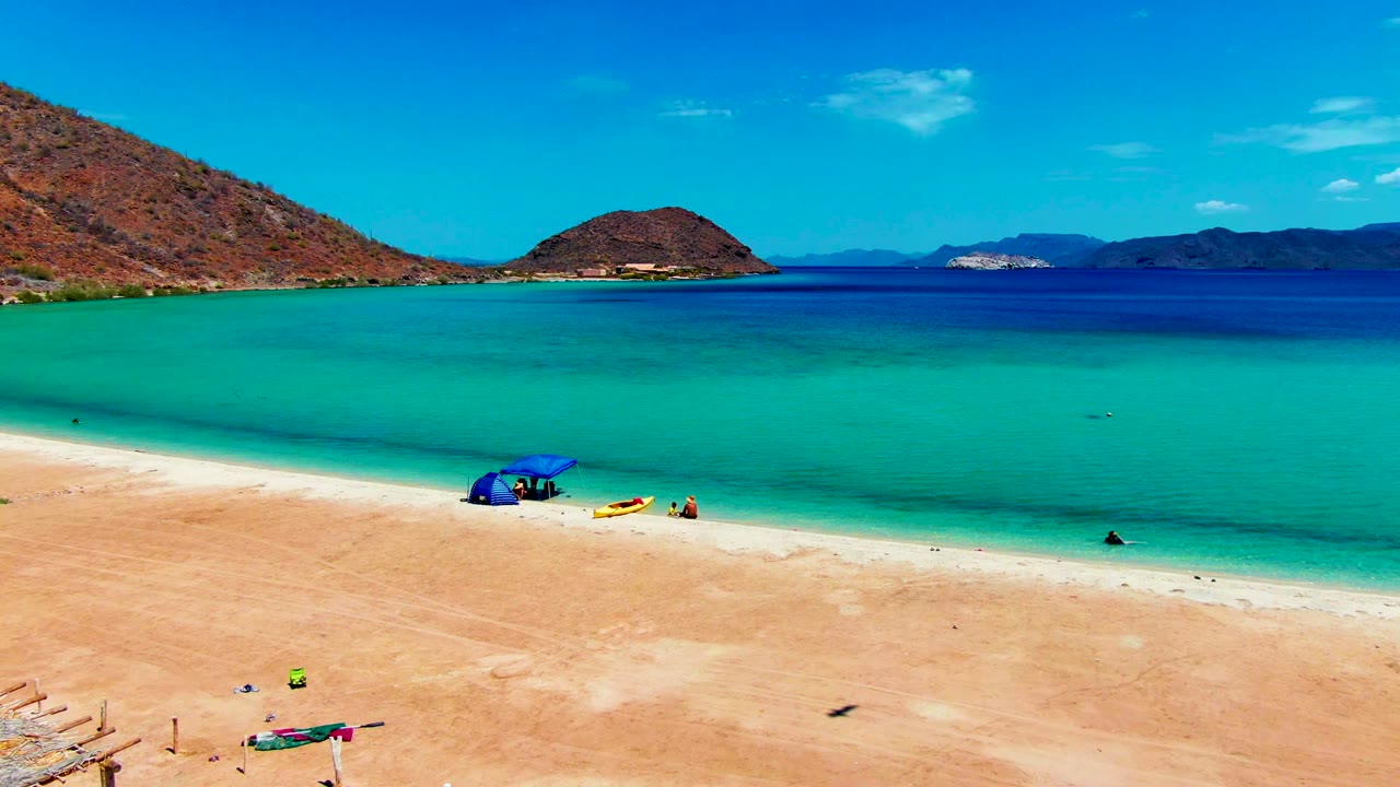 At 0:02, Playa Santispac fills the frame with pale sand in front, a blue umbrella and small beachgoers near the waterline, turquoise bay water, and low hills on the horizon.
