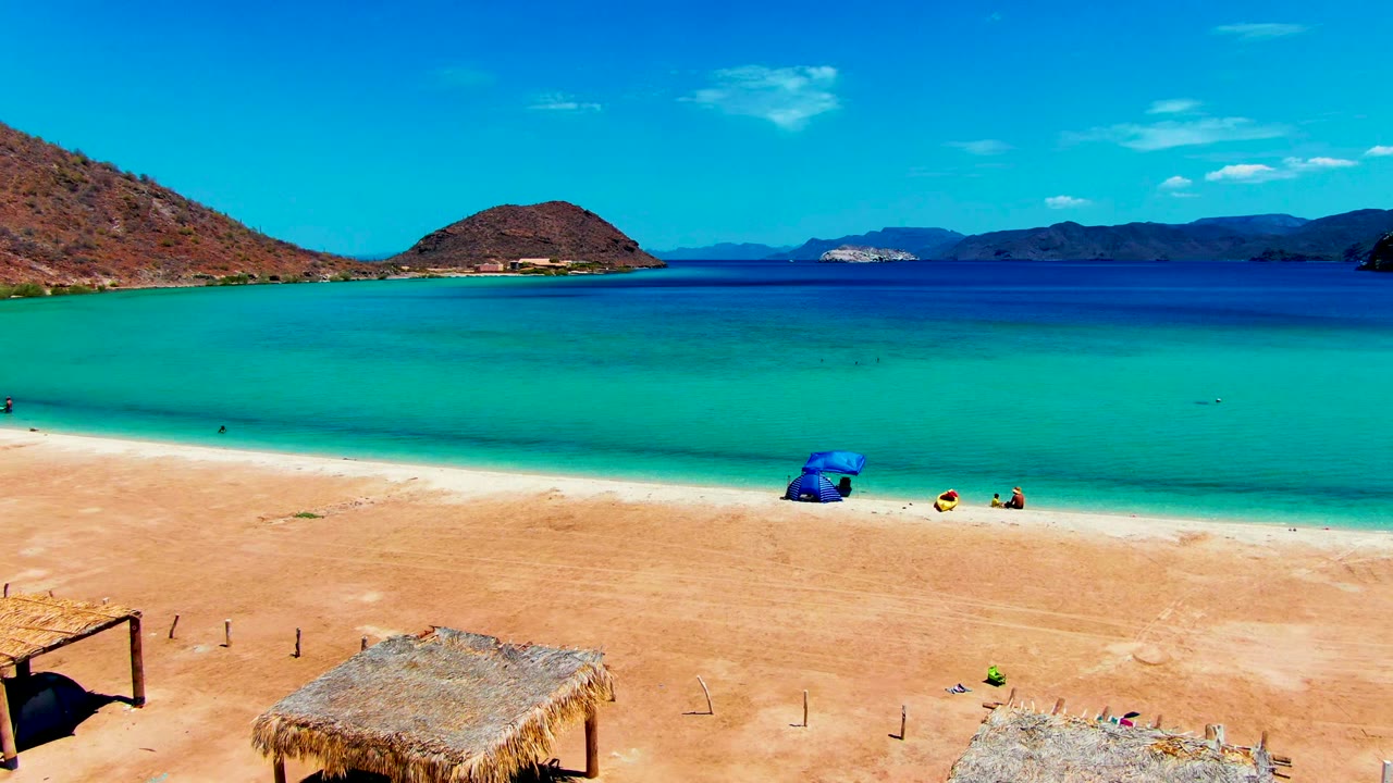 Opening frame of Playa Santispac, Baja California Sur, with thatched huts at the bottom edge, a blue umbrella near the shoreline, tiny beachgoers, turquoise water, and mountain ridges beyond.