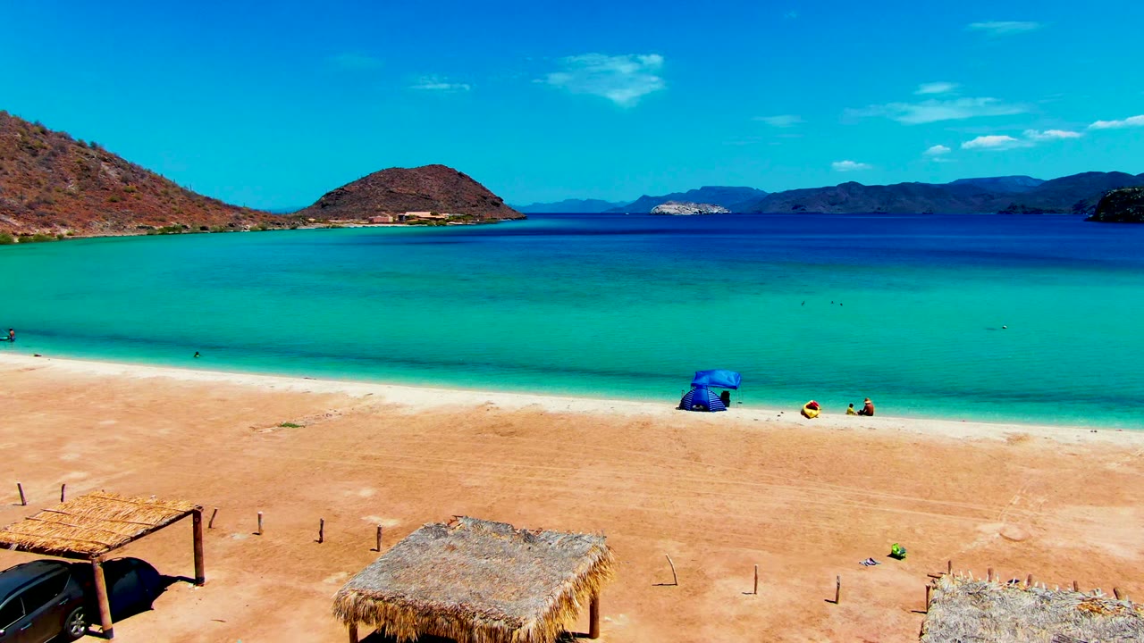 At 0:01 in Playa Santispac, the aerial view includes a dark car partly visible below a thatched shade roof, a blue umbrella by the water, small people on the sand, and low mountains across the bay.