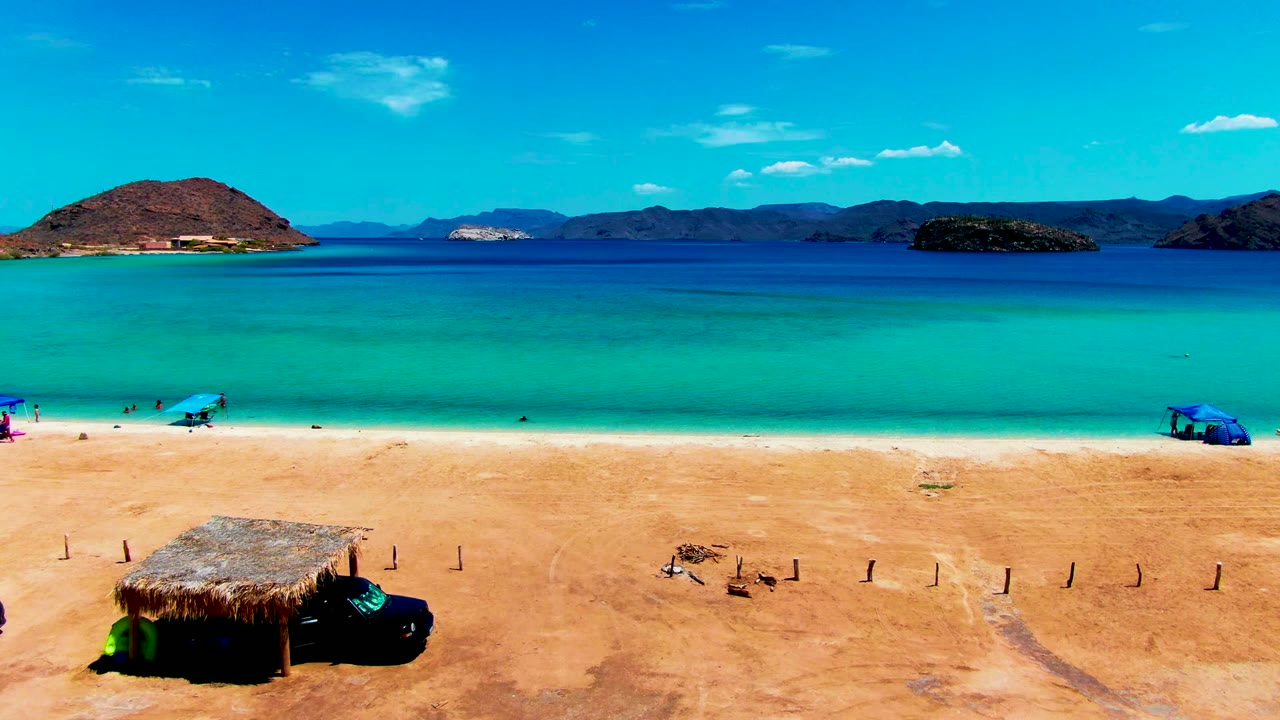 Playa Santispac beach view with a thatched hut and dark vehicle at the lower left, turquoise water across the middle, distant mountains, blue umbrellas, and small people visible near the shoreline.