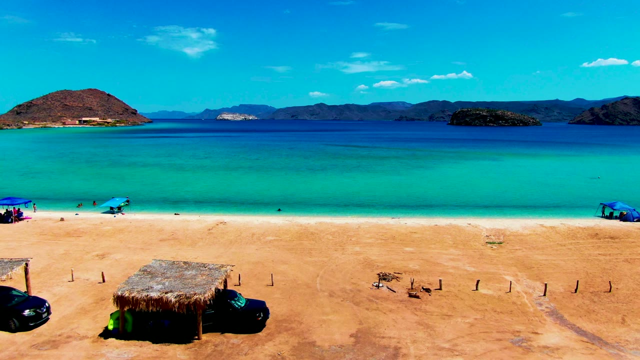 Thatched hut on sandy beach below turquoise water and distant mountains.