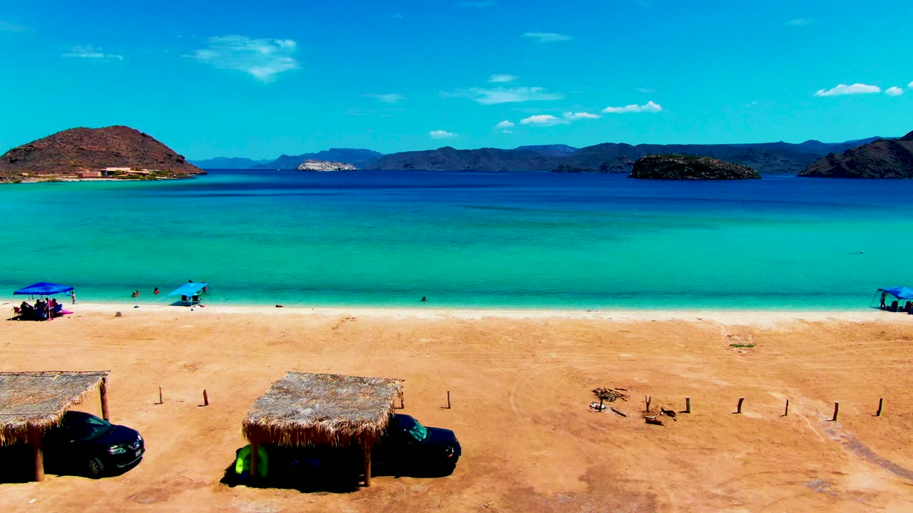 Playa Santispac beach frame with blue umbrellas and small people along the white shoreline, a thatched hut and vehicle in the sandy foreground, turquoise water, distant islands or mountains, and clear sky.