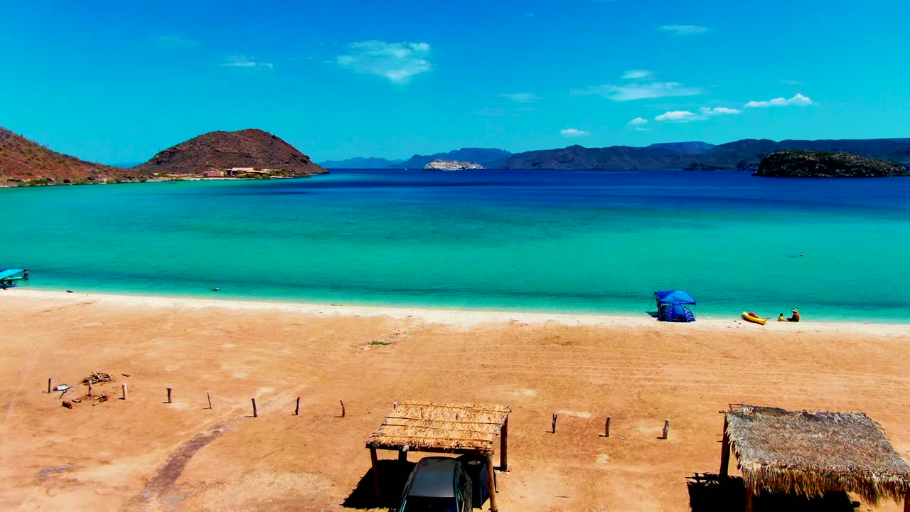 Opening view of Playa Santispac with sandy foreground, thatched hut roofs at the bottom edge, turquoise water, distant hills, and no recognizable people visible.
