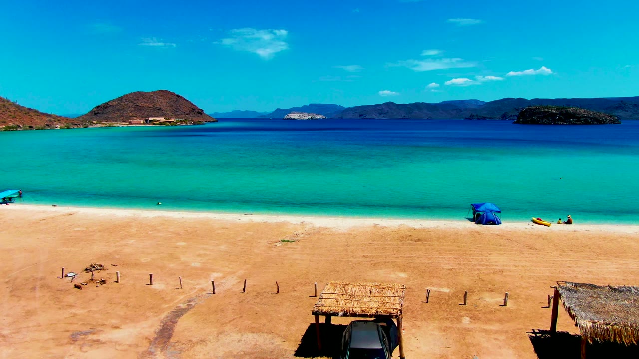Playa Santispac shoreline with a blue canopy near the water, a yellow watercraft on the sand, thatched hut roofs below, and no recognizable people visible.