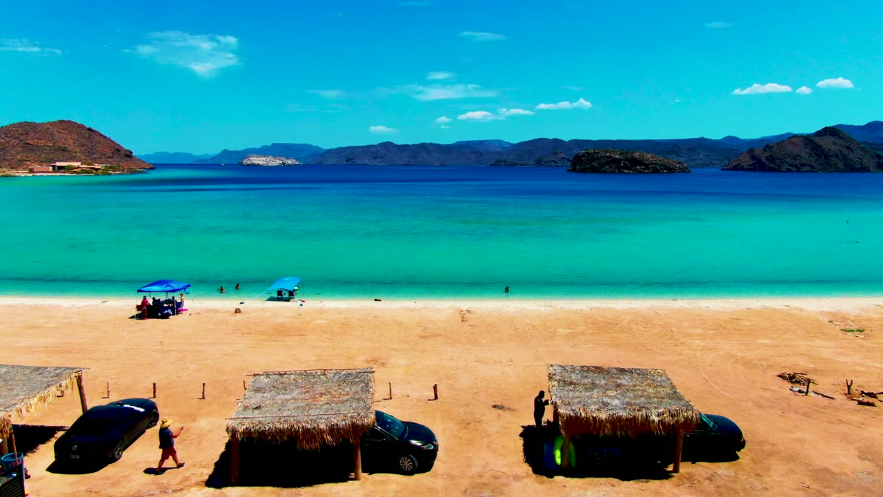 At 0:00, Playa Santispac appears from an elevated angle with thatched huts and cars along the sand, small people near the shoreline, turquoise water, and dark coastal hills beyond the bay.