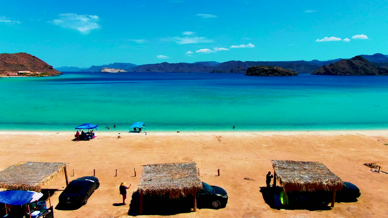 Beach huts and cars below clear turquoise water with swimmers near shore.