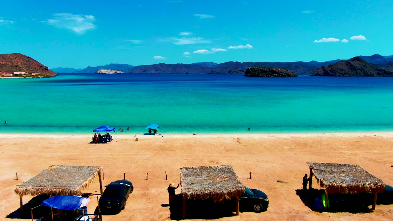 At 0:02, Playa Santispac remains framed with two thatched huts near the bottom edge, dark vehicles beside them, tiny people on the sand and in the water, and low mountains across the bay.