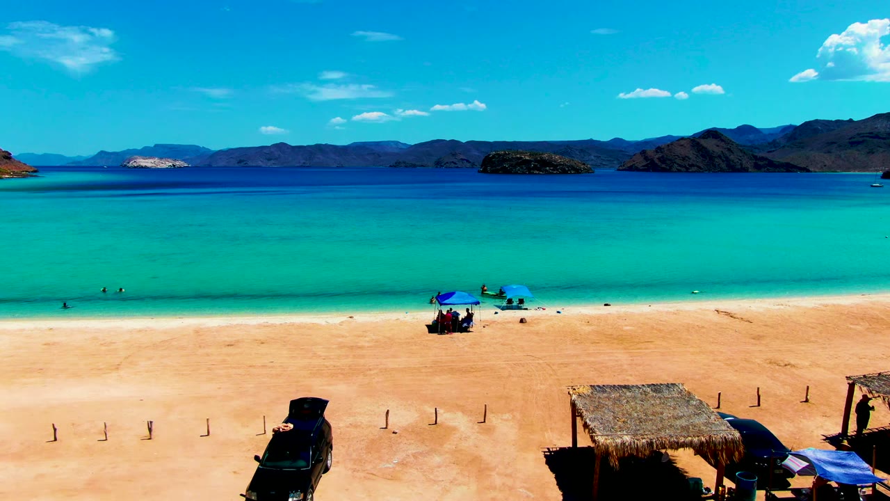 Opening frame of Playa Santispac with a dark vehicle at the lower left, blue umbrellas and tiny beachgoers near the shoreline, turquoise water, distant mountains, and a thatched shade structure at the lower right.