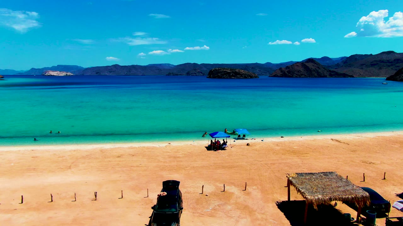 Vehicle on sandy beach below turquoise water with umbrellas, people, huts, and mountains.