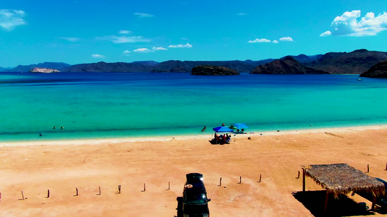 At 0:02, Playa Santispac remains in a wide aerial-style view, with the dark vehicle near the lower center, a thatched shade structure on the right, umbrellas and tiny figures at the surf line, and mountains across the bay.