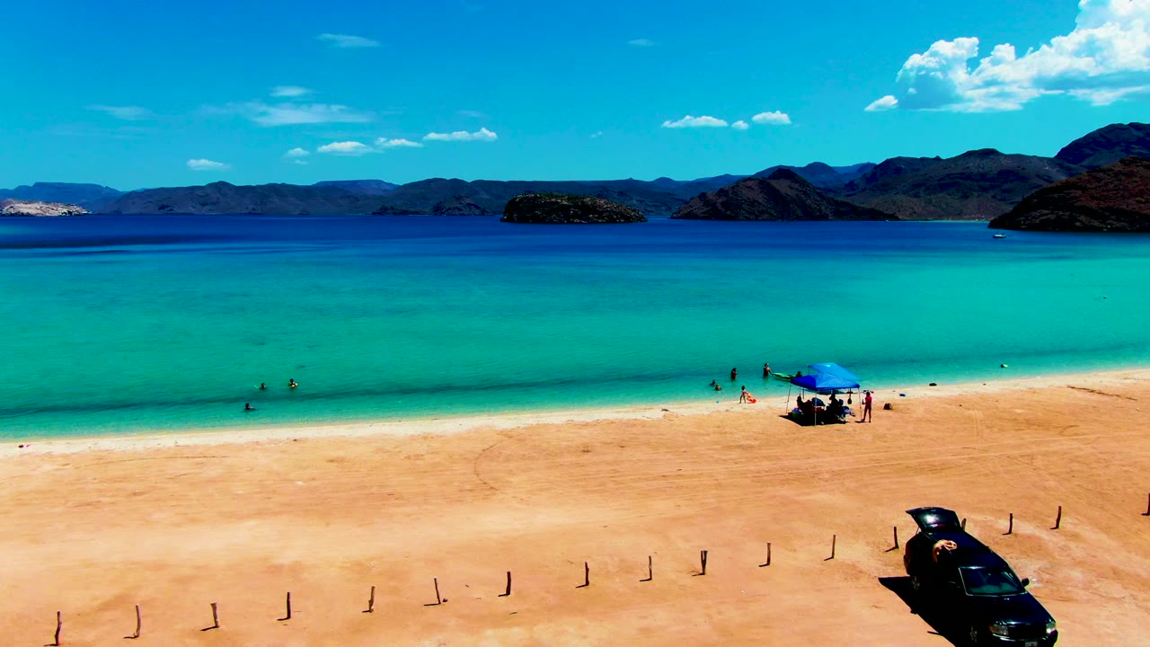 At Playa Santispac, a wide elevated frame shows turquoise shallows, small swimmers near the shoreline, a blue tent on the sand, a black car in the lower right, and mountains beyond the bay.