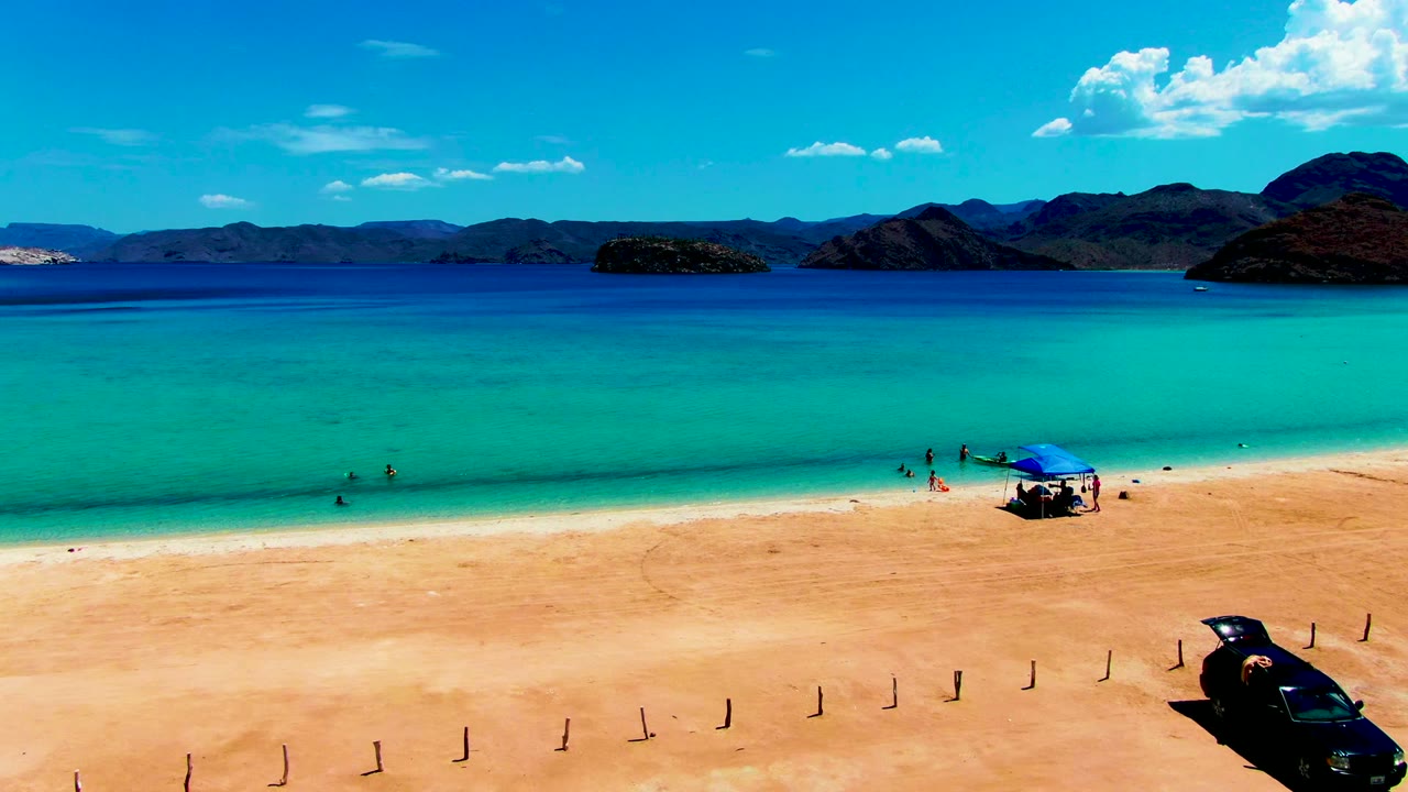 Aerial beach view with swimmers in clear water, a blue tent near the shore, and a black car on the sand.