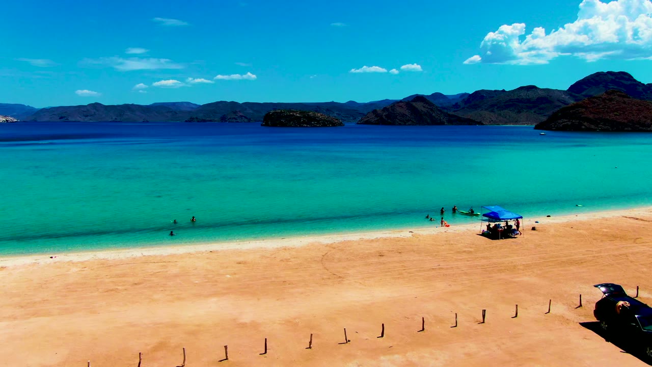 At 0:02 in Playa Santispac, the blue tent and beachgoers sit farther right in the frame, with the black car partly near the lower-right edge and the turquoise shallows stretching across the foreground waterline.