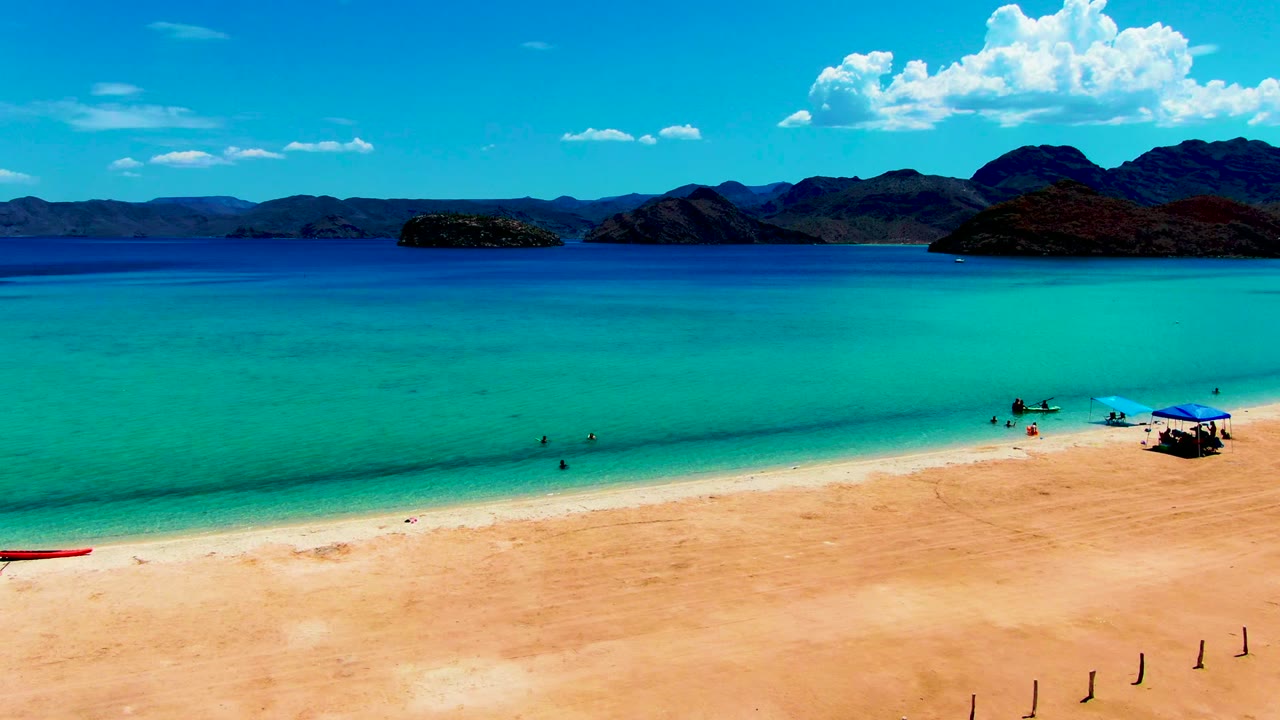 Opening frame of Playa Santispac with a red boat on the sand, small people wading in turquoise water, a blue umbrella at right, and dark mountains across the horizon.