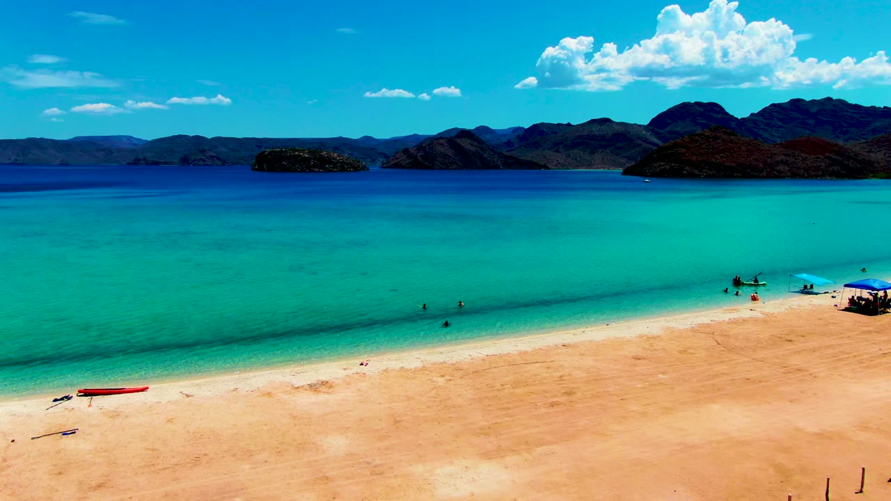 Later frame at Playa Santispac showing broad turquoise shallows, several small wading figures near center and right, the red boat on the sand, and dark mountains beneath scattered clouds.