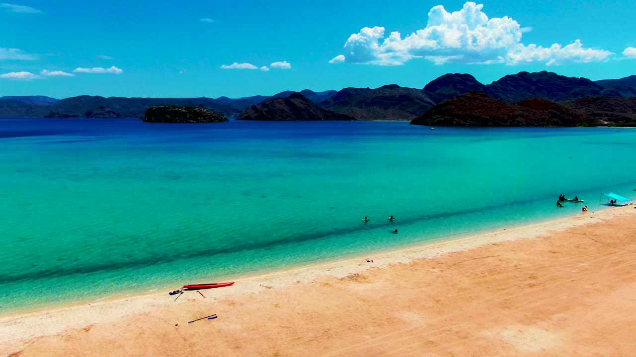 At Playa Santispac, pale sand fills the lower frame while clear turquoise water holds small swimmers near shore, with a red boat on the beach and dark mountains under scattered clouds.
