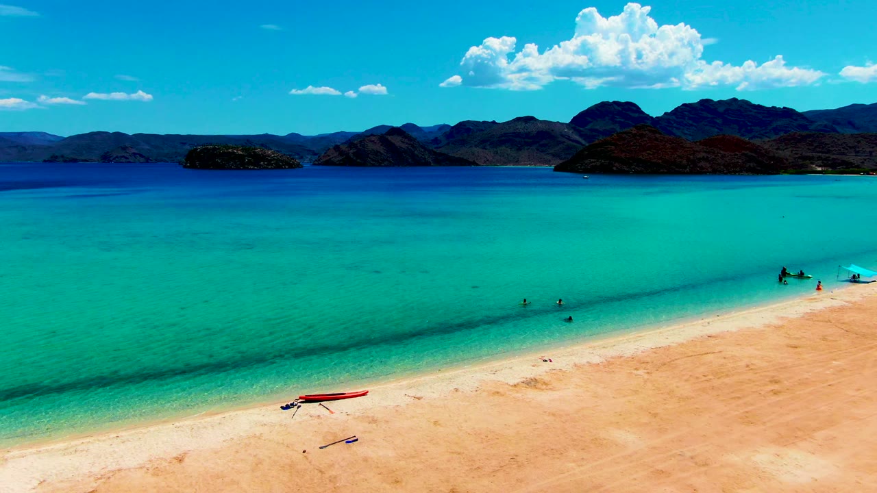 Clear shallow water beside sand with swimmers, a red boat, mountains, and clouds.
