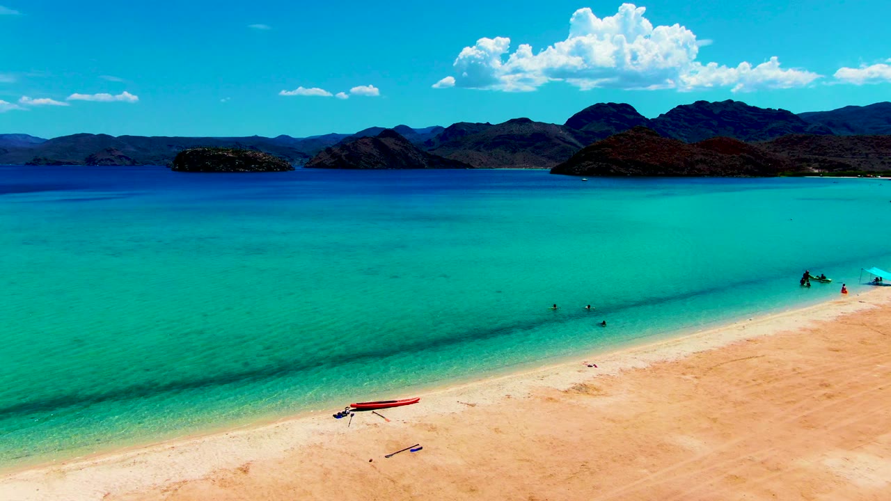 At 0:02, the frame shows more open turquoise water between the sandy foreground and the mountain horizon, with small people wading near the shore and the red boat still visible on the beach.