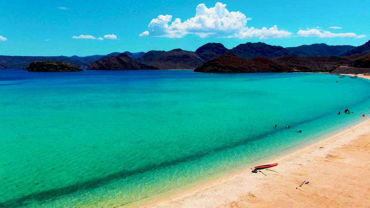At 0:00, Playa Santispac is seen from above with clear turquoise shallows, people wading near the beach, a red boat on pale sand, and dark mountains under a blue sky.