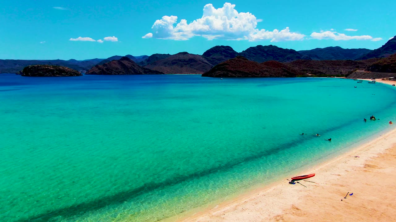Turquoise bay with people near shore, a red boat on sand, and mountains beyond.