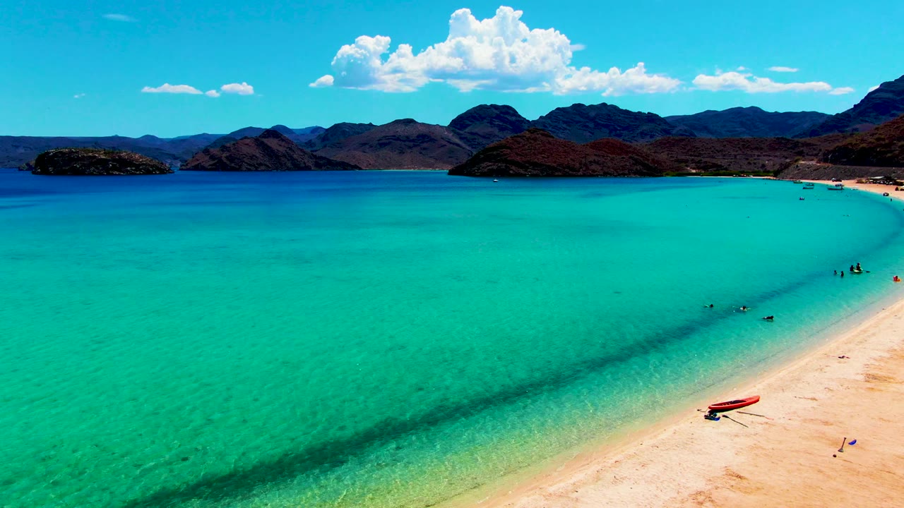 At 0:02, Playa Santispac fills the frame with broad turquoise water, a diagonal sandy shoreline, visible people in the shallows, a red boat on the sand, and bright clouds above the mountains.
