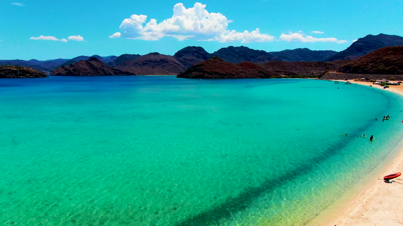 Aerial frame from Playa Santispac showing turquoise shallows across the foreground, a sandy beach at right with small visible people, dark mountains on the horizon, and white clouds in a blue sky.