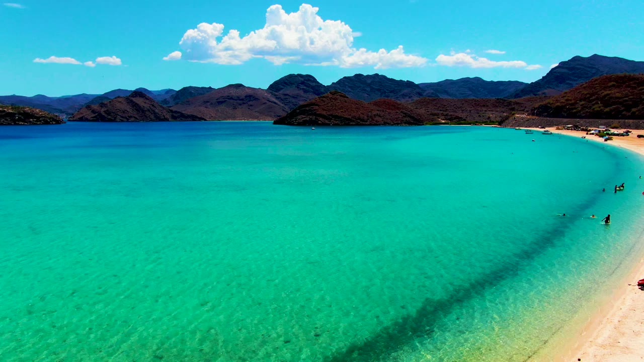 Turquoise shallows beside a sandy shoreline with swimmers and distant mountains.
