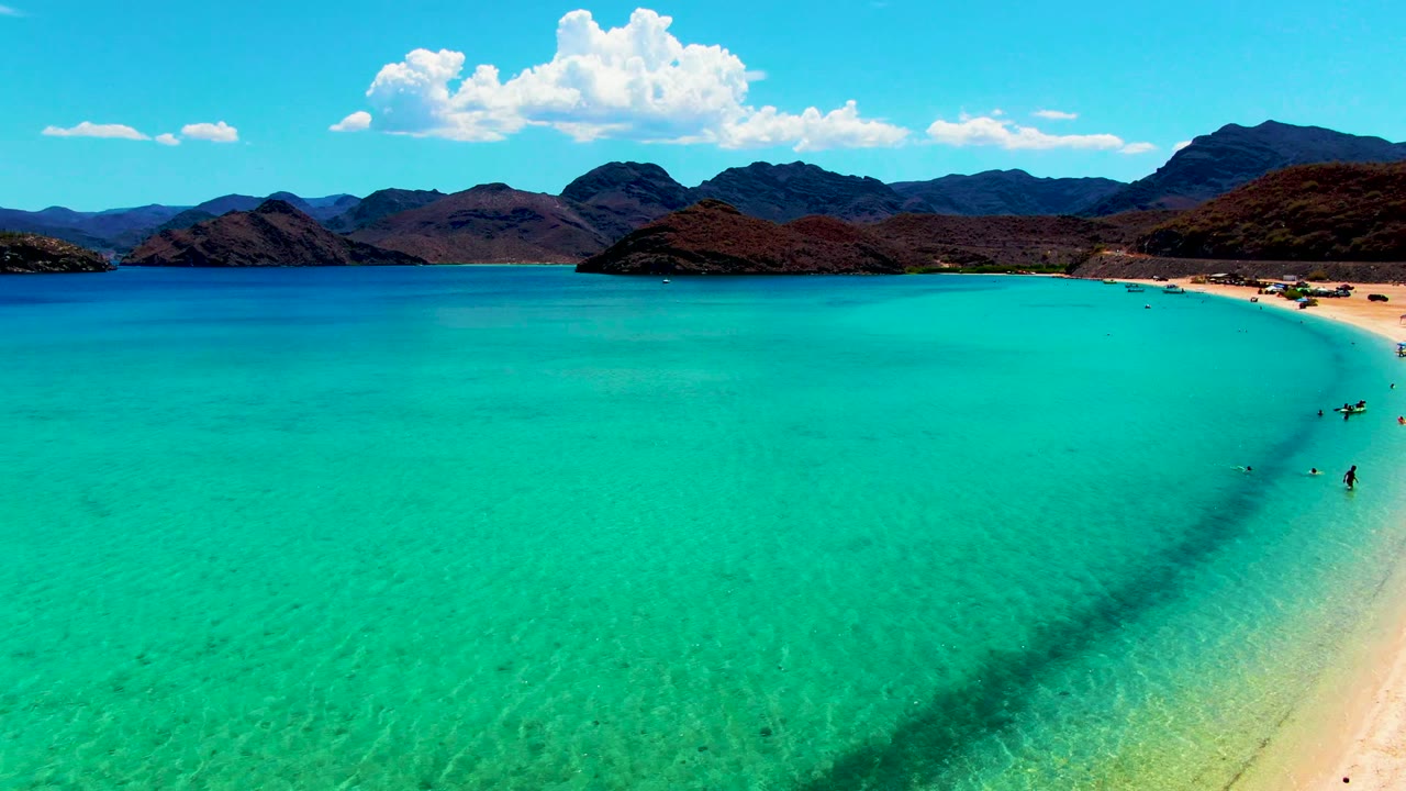 At 0:02, the aerial view holds on clear turquoise water with faint seabed texture, visible people clustered near the right-side shoreline, pale sand, distant mountains, and a bright cloud above the horizon.