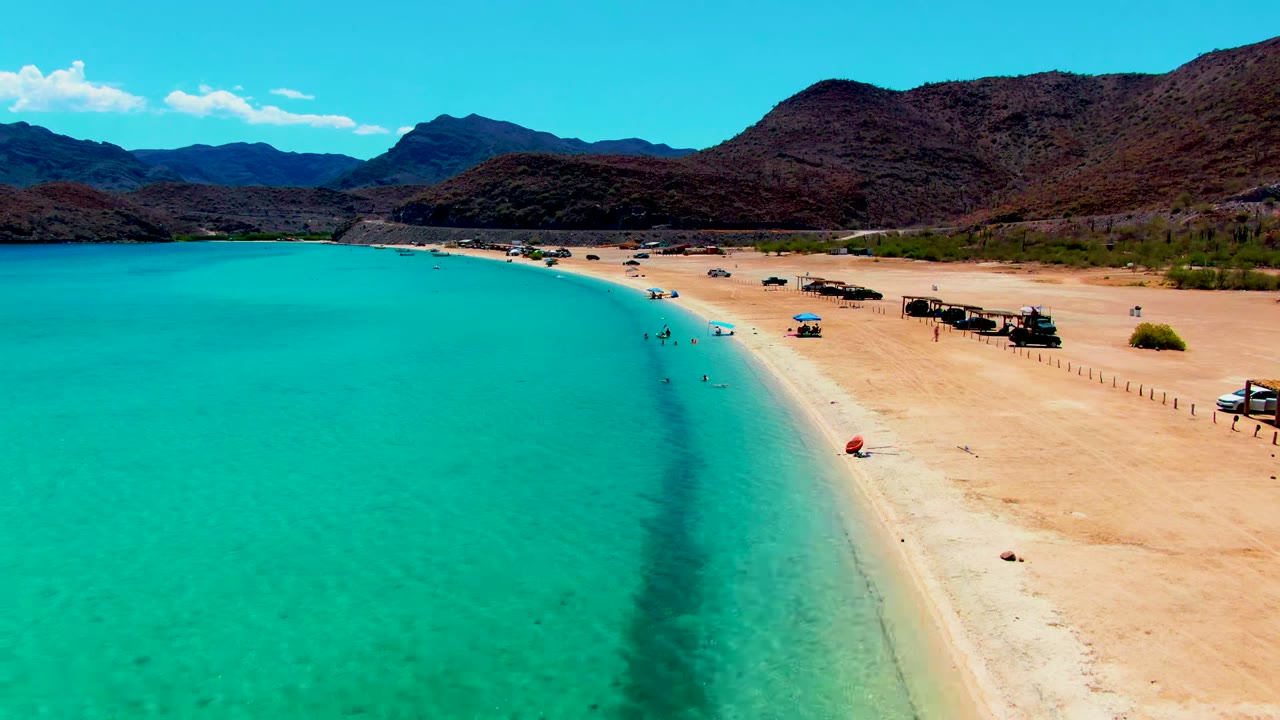 Aerial view over Playa Santispac with turquoise water on the left, a sandy shoreline on the right, small beachgoers near the water, parked vehicles, shade huts, umbrellas, and mountains beyond.