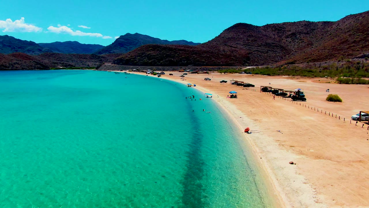 Wide aerial beach view with clear turquoise water, swimmers near shore, parked vehicles, huts, and mountains.