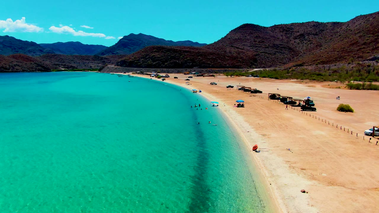 High view of Playa Santispac with the curved waterline running through the frame, small figures in the water, an orange object near the beach edge, vehicles and huts on the sand, and mountains under clear light.