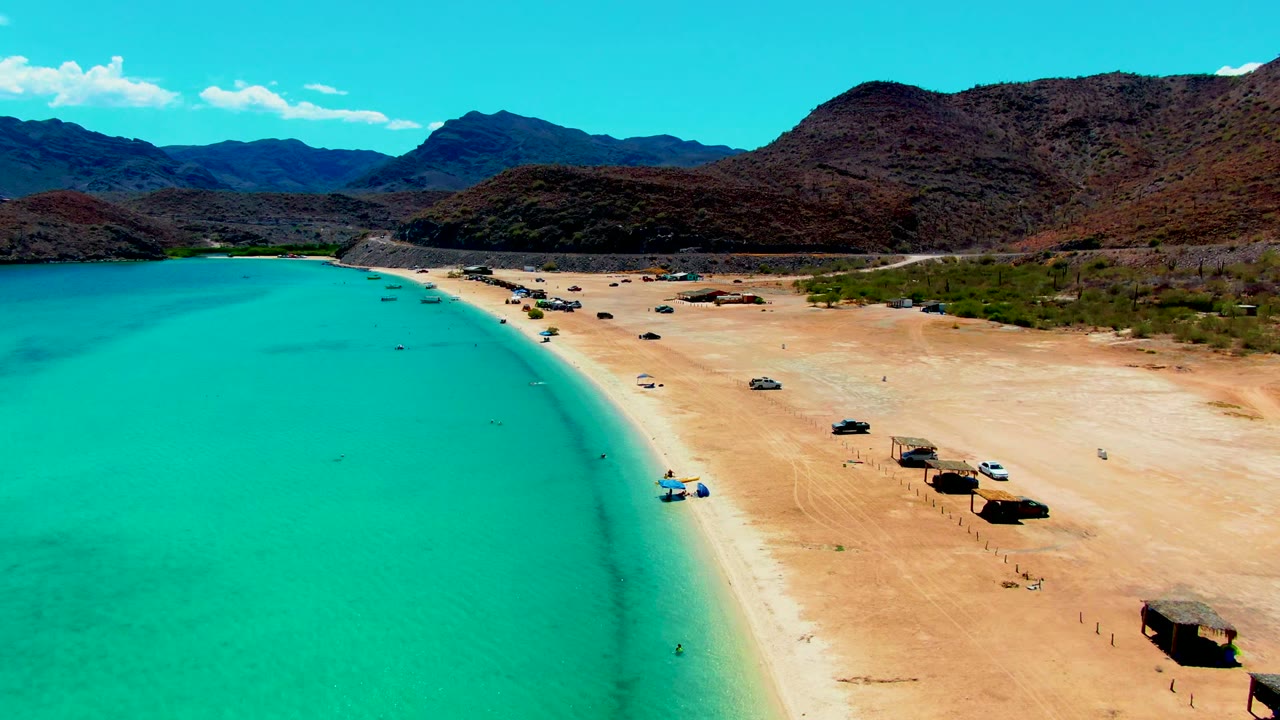 Aerial frame over Playa Santispac showing turquoise water at left, a sandy beach with scattered vehicles and shade huts, arid mountains beyond, and no visible people.