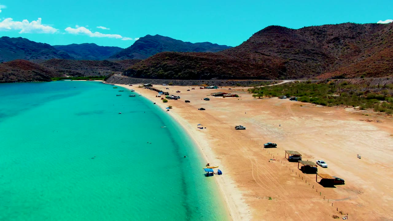 Aerial view at Playa Santispac with the curved turquoise shoreline closer to frame center, parked vehicles on the sand, small shade huts near the waterline, mountains in back, and no visible people.