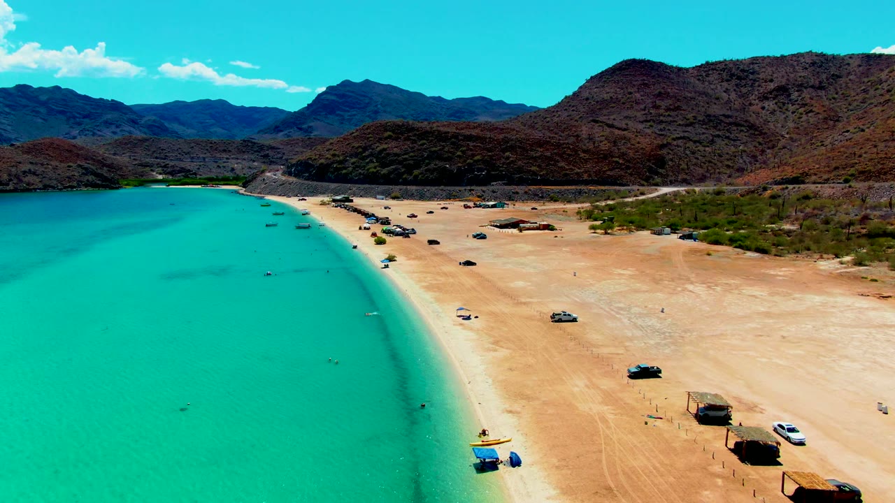 Wide drone frame of Playa Santispac showing bright turquoise water, broad sand marked by vehicle tracks, several beach vehicles, thatched huts, desert hills, and no visible people.
