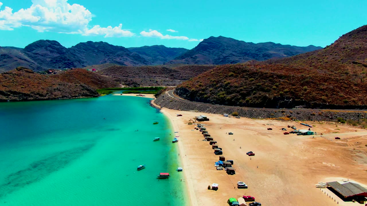 Opening aerial frame over Playa Santispac shows turquoise water beside a sandy beach with small boats, rows of vehicles, desert hills, distant mountains, and no visible people.