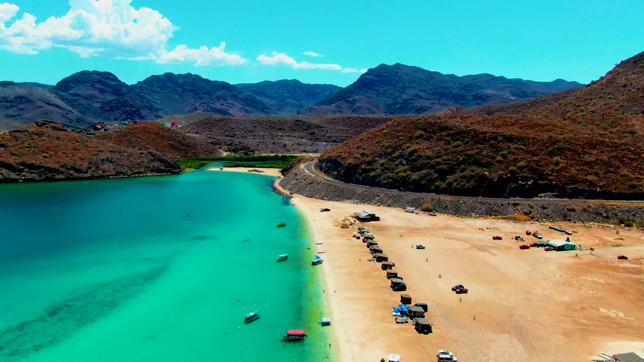 Aerial shoreline with boats near turquoise water and vehicles on sand.