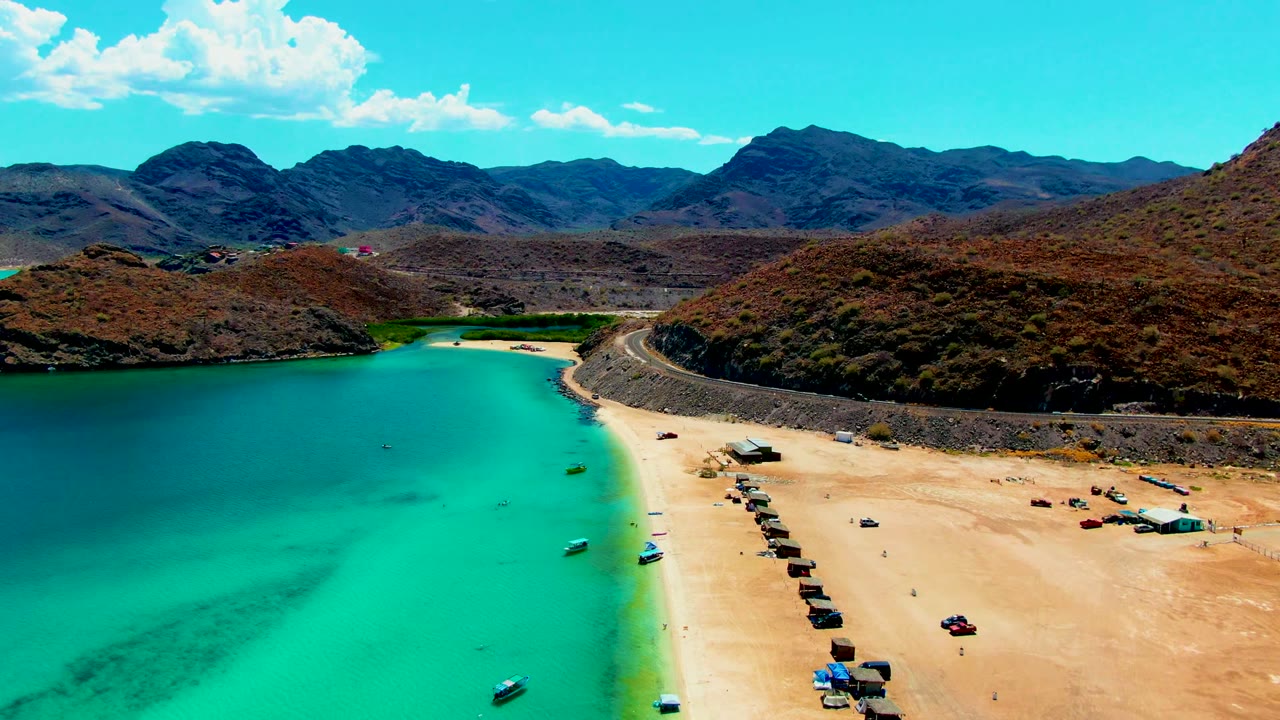 At 0:02, the aerial view emphasizes the road tracing the beach at Playa Santispac, with turquoise water, several small boats, scattered vehicles, rocky hills, and no visible people.