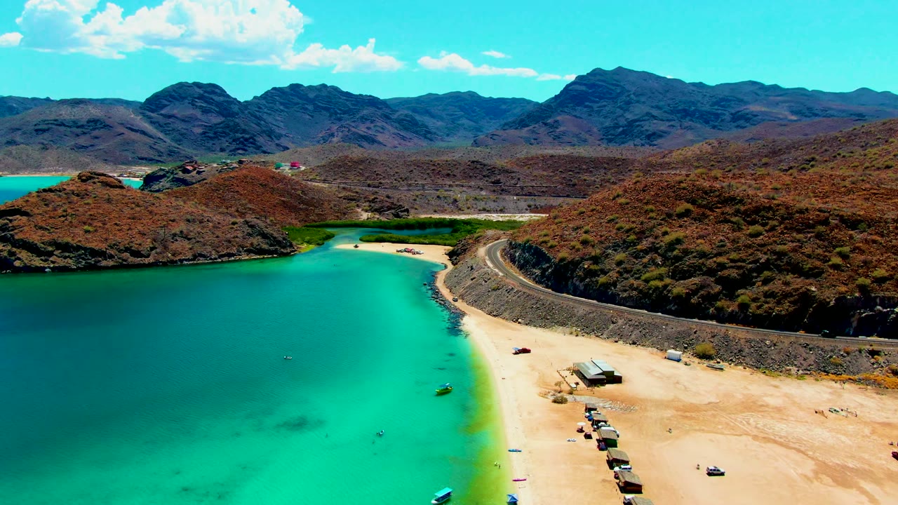 Aerial frame over Playa Santispac shows turquoise water curving around a sandy beach, a road below dry hills, distant mountains, scattered clouds, and no visible people.