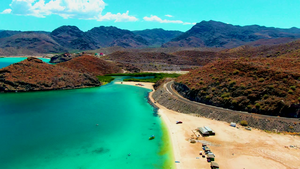 Turquoise bay and pale beach beside a curving road with desert hills behind.
