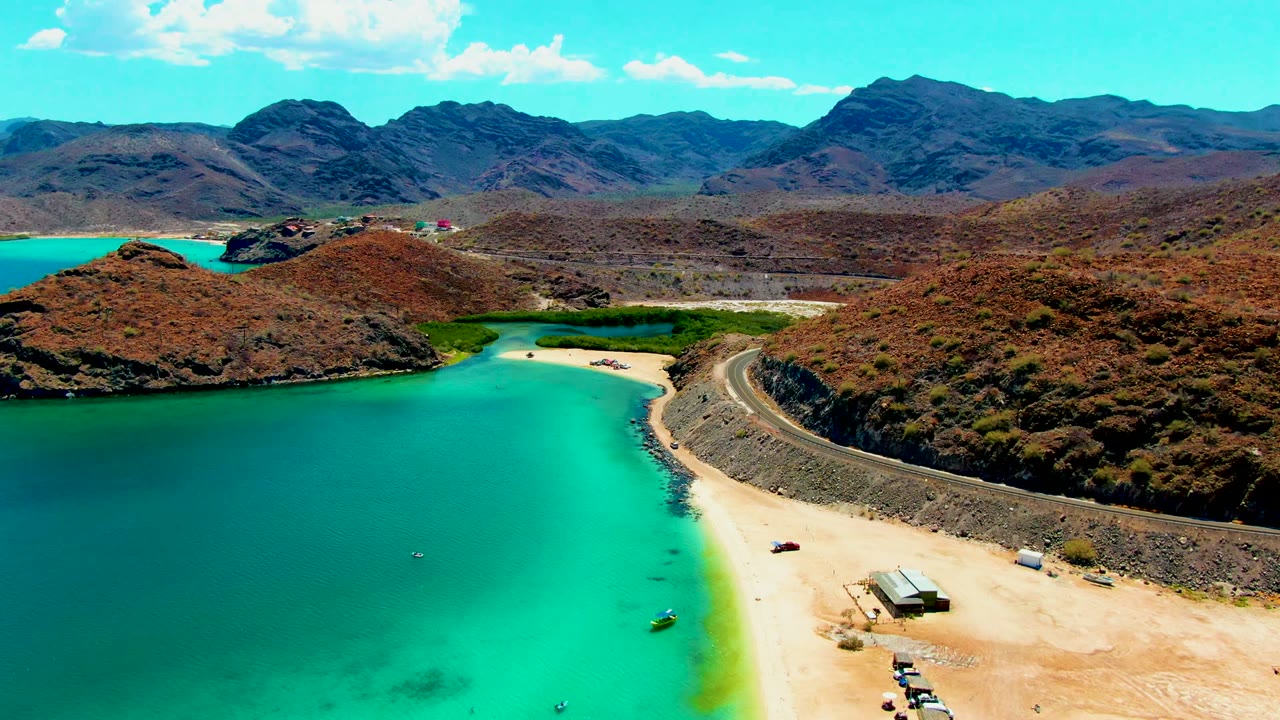 At Playa Santispac, the drone view shows a broad turquoise inlet, beach access area, hillside road, arid ridges, distant mountains, and no visible people.