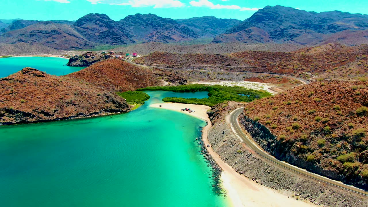 Opening aerial frame at Playa Santispac shows turquoise water beside a pale beach, a curving coastal road, dry hills, distant mountains, clear sky, and no visible people.