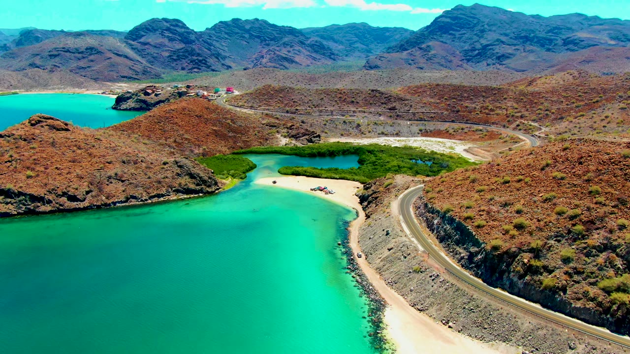 Turquoise shoreline and coastal road curve around dry hills under a clear sky.