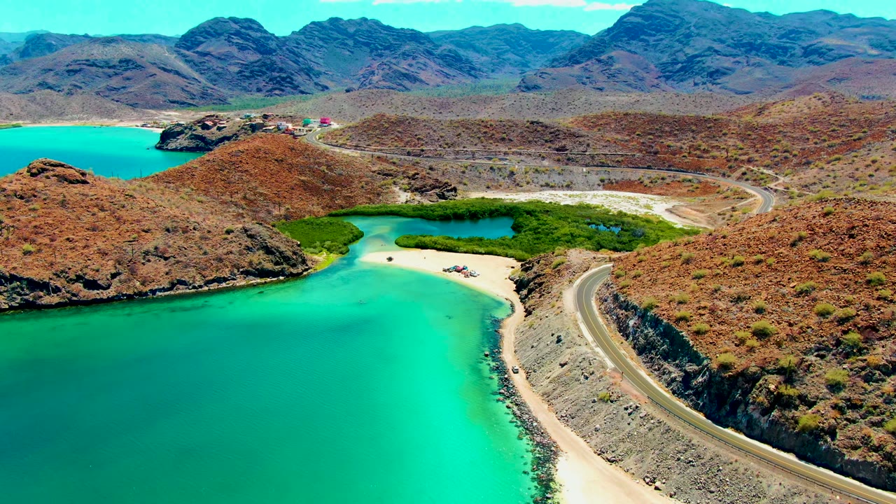 At 0:02, Playa Santispac appears from above with more open turquoise water in the foreground, the road along the right slope, dry hills beyond, and no visible people.