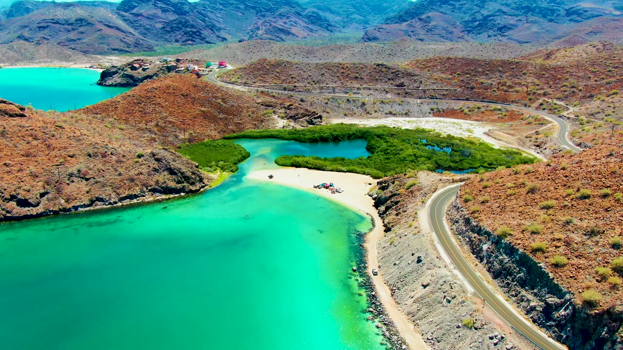 Aerial frame at Playa Santispac, Baja California Sur, showing turquoise water in the foreground, a sandy beach beside a green lagoon, rocky hills, a curving coastal road, distant mountains, and no visible people.