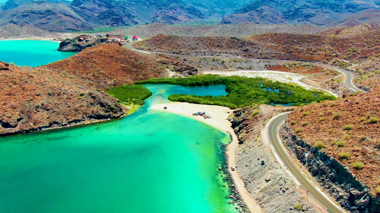 Aerial view of a coastal road beside bright turquoise water and a small beach with a green lagoon behind it.