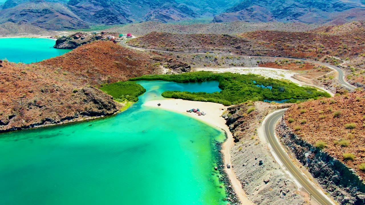 At 0:02, Playa Santispac appears from above with the road curving along the right edge, turquoise bay water across the lower frame, the green lagoon centered, dry rocky hills beyond, and no visible people.